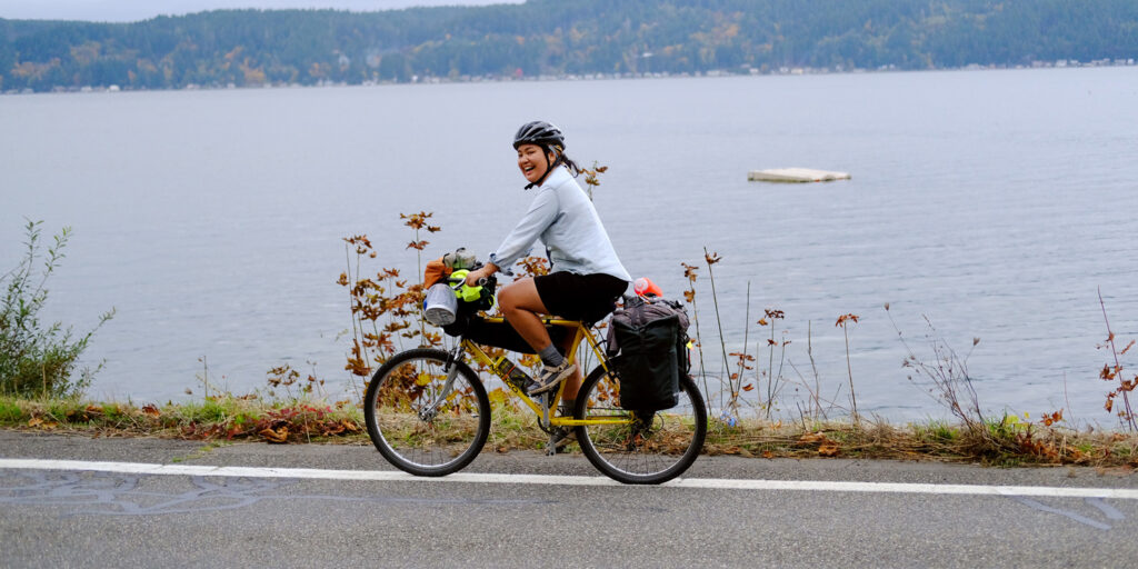 Person riding a loaded bike on a paved road past a large body of water.