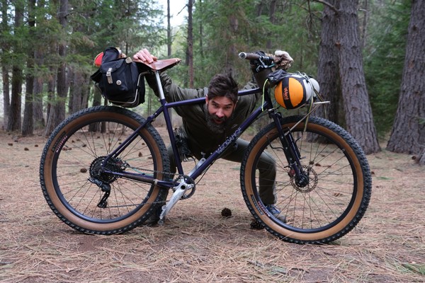 Smiling man with beard crouched with his head in the triangle of his loaded bikepacking set up.
