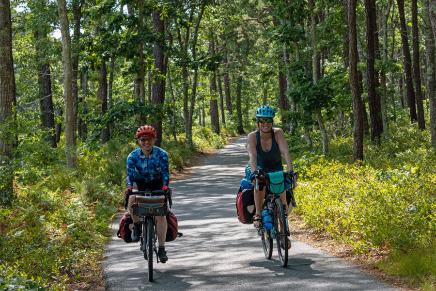  Two smiling people ride on a paved trail in the woods.
