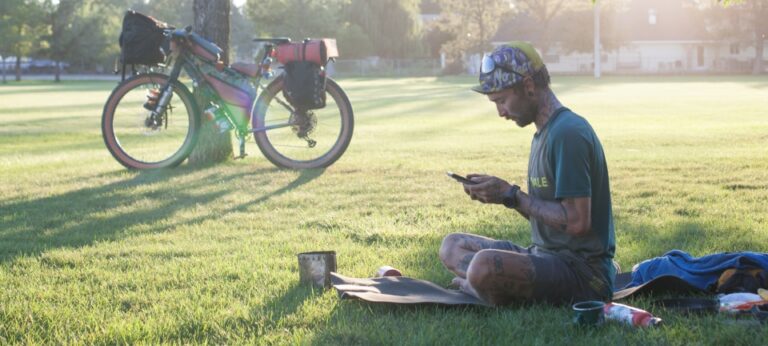 A cyclist rests in a city park at sunset, his gravel bike rests against a tree in the background